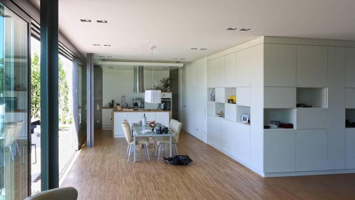 Offene Wohnküche mit modernem Design Heller Wohnbereich mit offener Küche, integriertem Essbereich, großen Fenstern zum Garten und einer weißen Einbauschrankwand mit offenen Fächern.